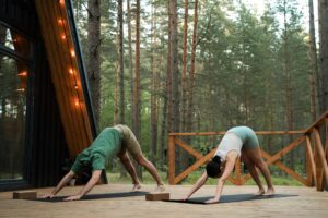 A couple performs yoga on a cabin deck nestled in a serene forest setting.