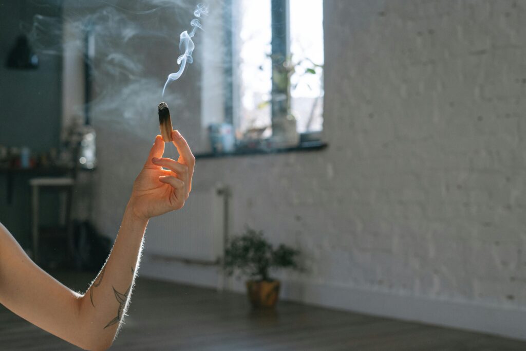 A man's hand holding a burning Palo Santo stick with smoke wafting in a sunlit room, creating a serene atmosphere.